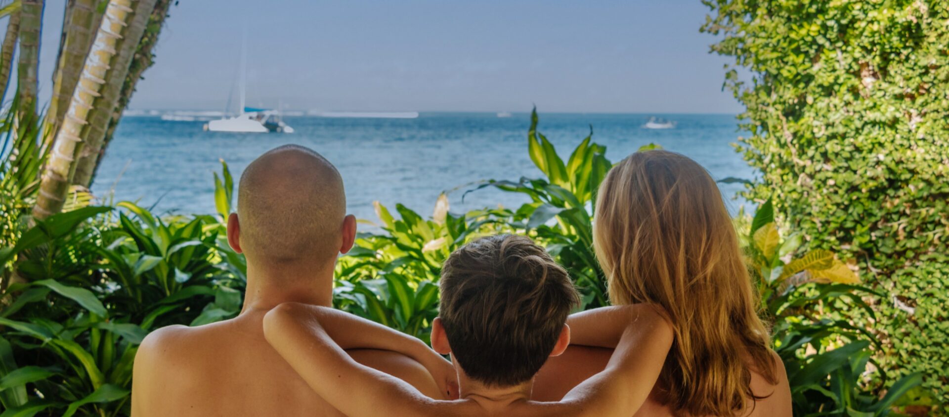 Famille de trois personnes de dos regardant la mer et un voilier depuis une cabane tropicale, entourée de palmiers et de verdure.