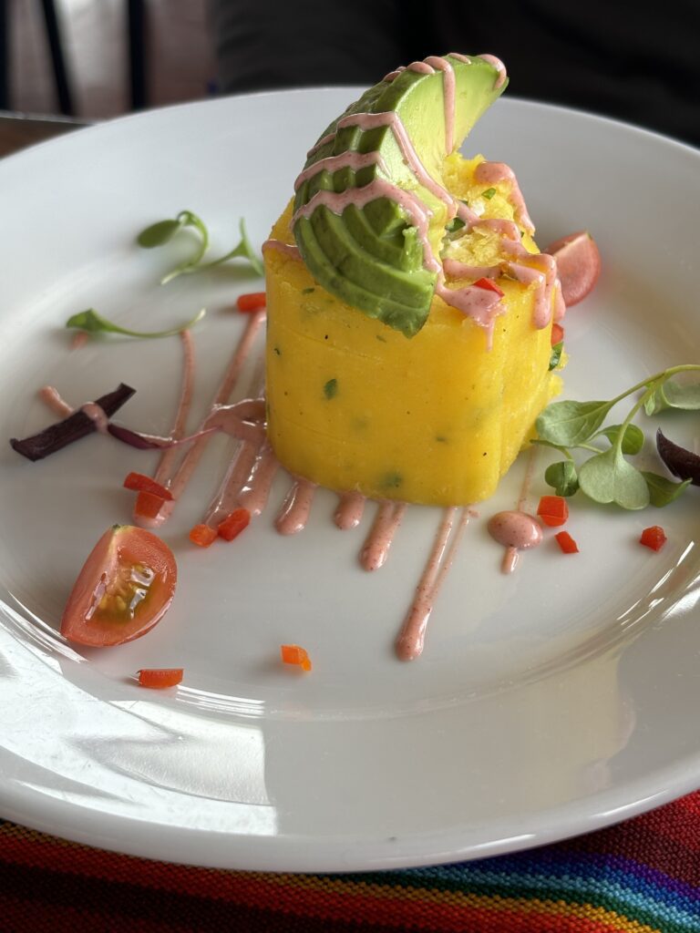 Assiette avec une causa péruvienne joliment dressée à base de purée de pommes de terre jaunes, avocat et sauce colorée.