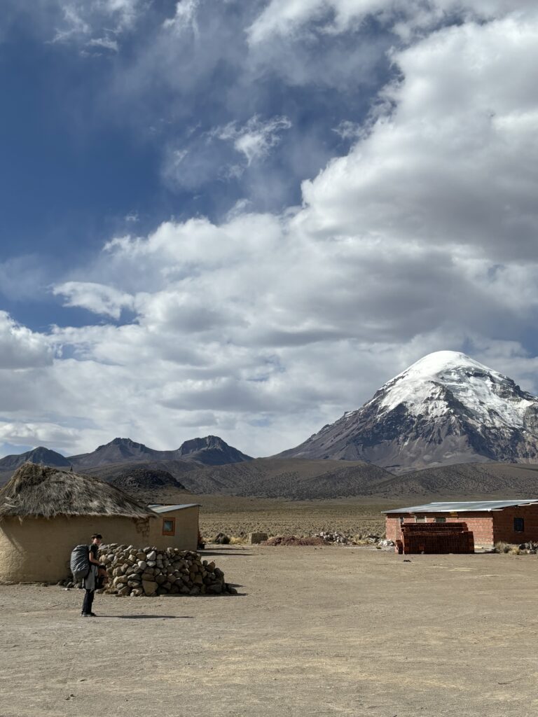 Paysage de montagne en Bolivie, avec des habitations traditionnelles en pierre et toit de chaume au premier plan, et un volcan enneigé à l’arrière-plan sous un ciel nuageux.