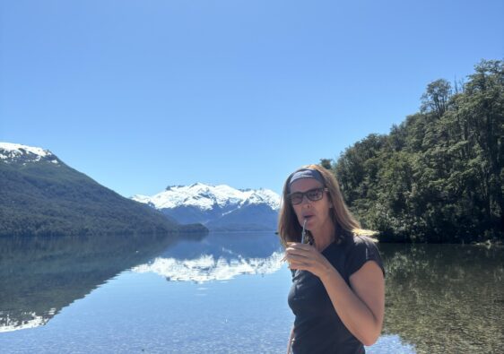 Une femme debout au bord d’un lac cristallin entouré de montagnes enneigées, dégustant du maté sous un grand ciel bleu en Patagonie.