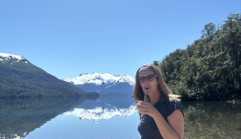 Une femme debout au bord d’un lac cristallin entouré de montagnes enneigées, dégustant du maté sous un grand ciel bleu en Patagonie.